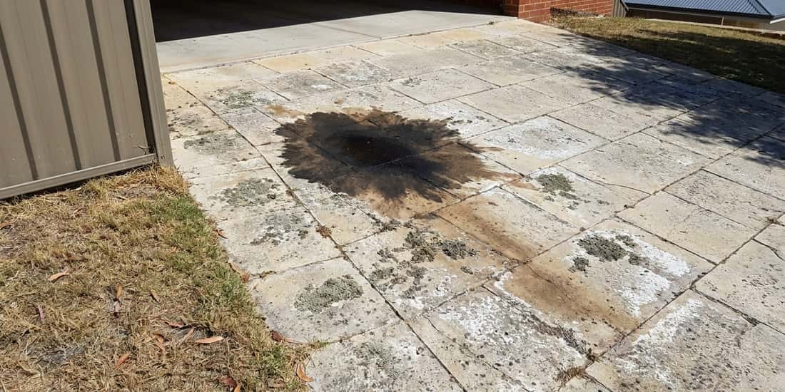 A dark oil stain spreads across limestone pavers in a driveway area, surrounded by patches of lichen and white mineral deposits, with a metal shed on the left and a brick house in the background.