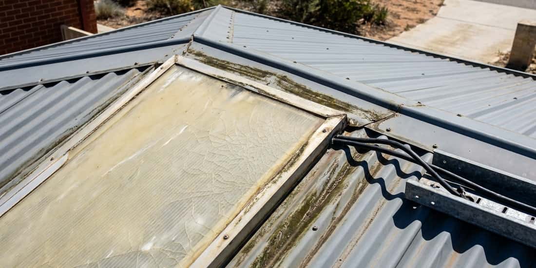 A weathered corrugated metal roof with a cracked, stained skylight panel and an unused solar panel mounting rail with cables running along the roof ridge.