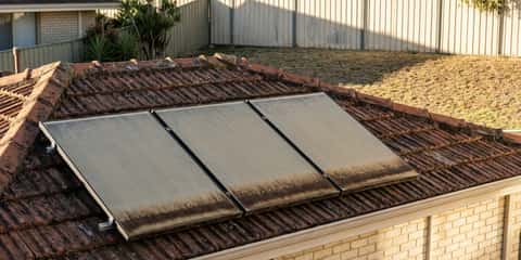 Three grimy solar water heating panels mounted on a red tile roof, with a brick house, corrugated steel fence, and lawn visible in the background.