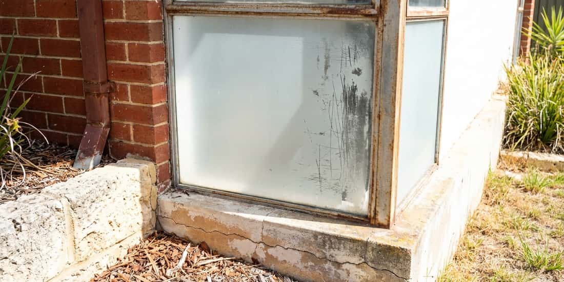 A corner window with frosted, streaked glass and a rusted frame sits above a cracked concrete sill on a brick house.
