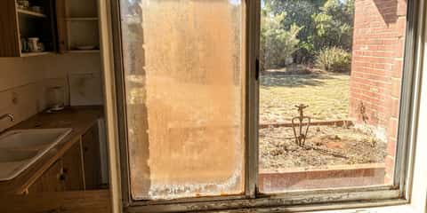 A grimy sliding glass window with heavy white mineral staining, viewed from a worn kitchen sink area, looking out onto a lawn with an old sprinkler and brick pillar.