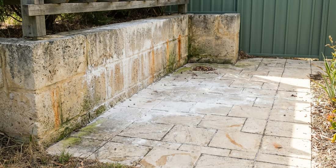 A paved patio with algae stains and rust marks sits beside a limestone retaining wall, wooden pergola, and green metal fence.