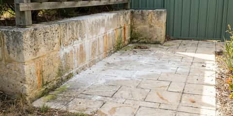 A paved patio with algae stains and rust marks sits beside a limestone retaining wall, wooden pergola, and green metal fence.