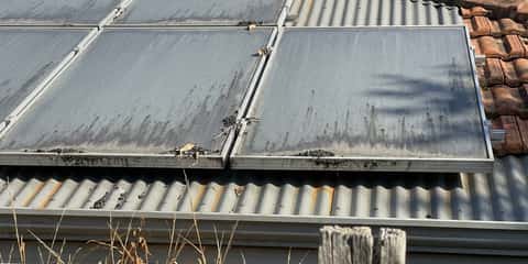Two rows of solar panels mounted on a tiled and corrugated metal roof, covered in dust and ash streaks with debris caught at the panel frame joints.