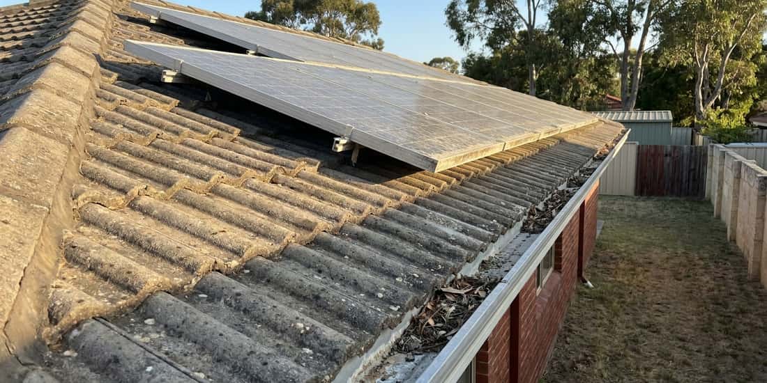 A weathered concrete tile roof on a brick house, with a tilted solar array mounted near the ridge and dry leaves collected in the gutter above a grassy backyard.