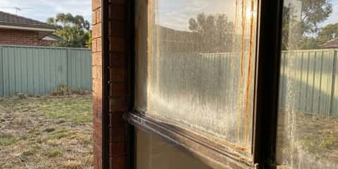 A dirty aluminium-framed window with rust streaks and white mineral buildup on the glass, viewed from beside a brick pillar with a backyard, colourbond fence, and trees visible in the background.
