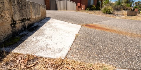 A driveway with exposed aggregate concrete meeting a plain concrete path, with rust-colored staining visible on the aggregate surface near a limestone wall.