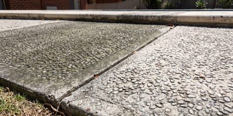 Close-up of two exposed aggregate concrete panels divided by a joint, with a brick house, fence and plants in the background.