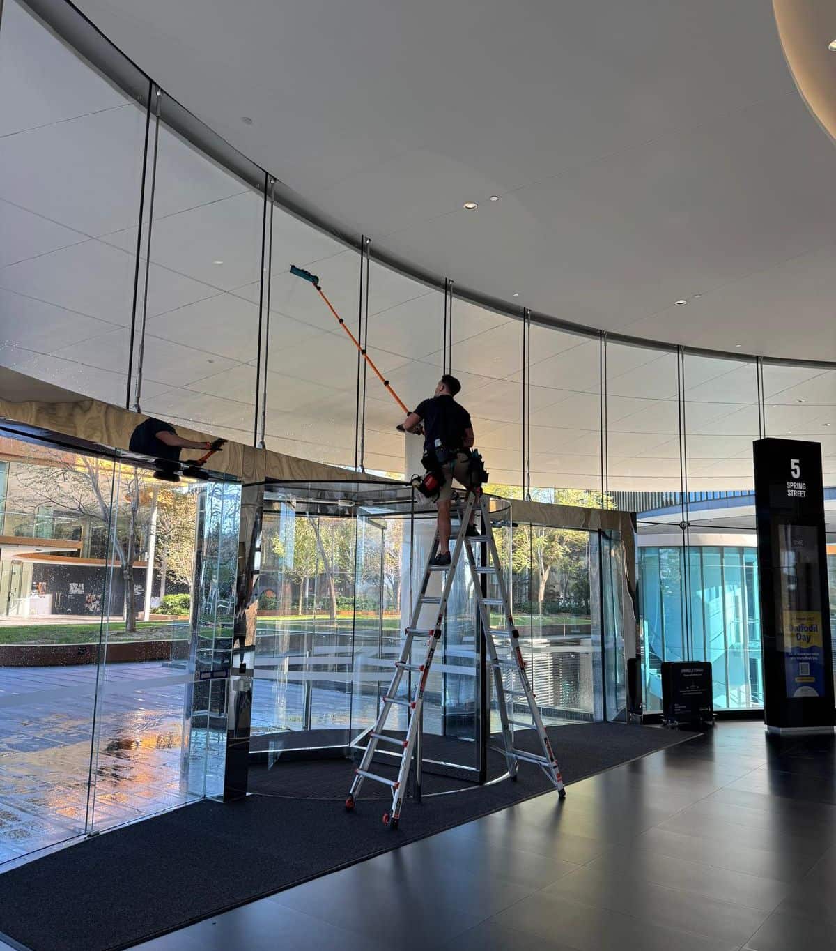 A Purified Group technician on a ladder pole-cleaning the curved glass atrium of a Perth commercial building entrance