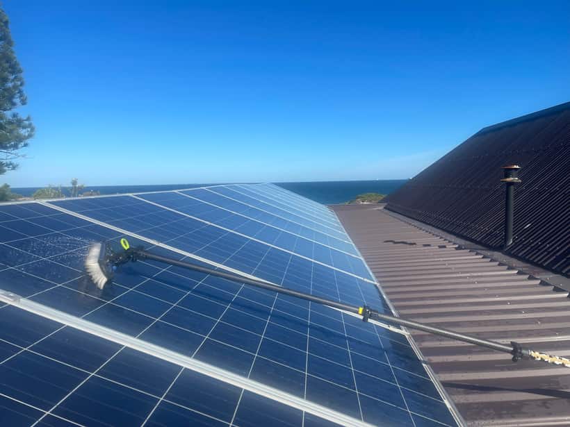 Purified water brush cleaning a rooftop solar array on a Perth home, the coast visible beyond the roofline