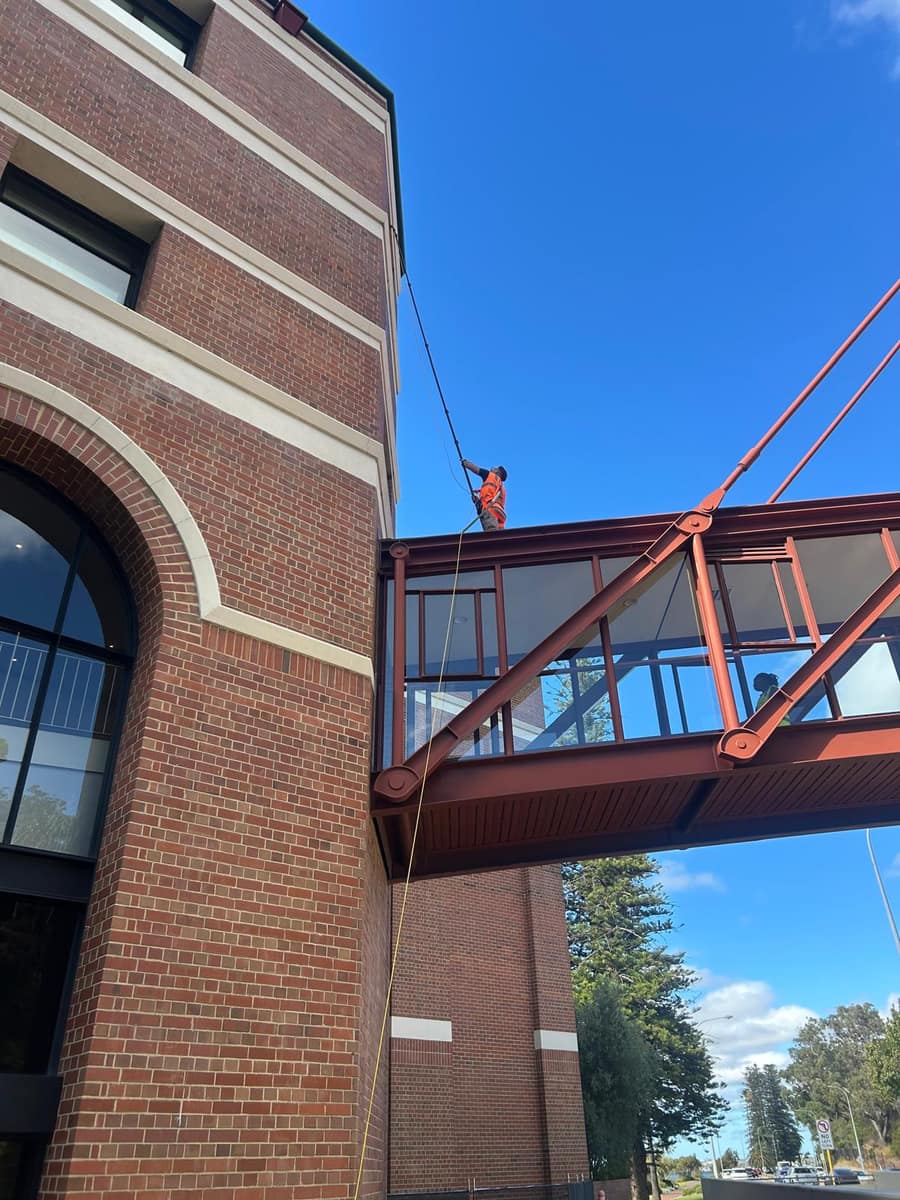 Technician on an elevated glass walkway pole-cleaning upper storey windows between heritage buildings, Perth