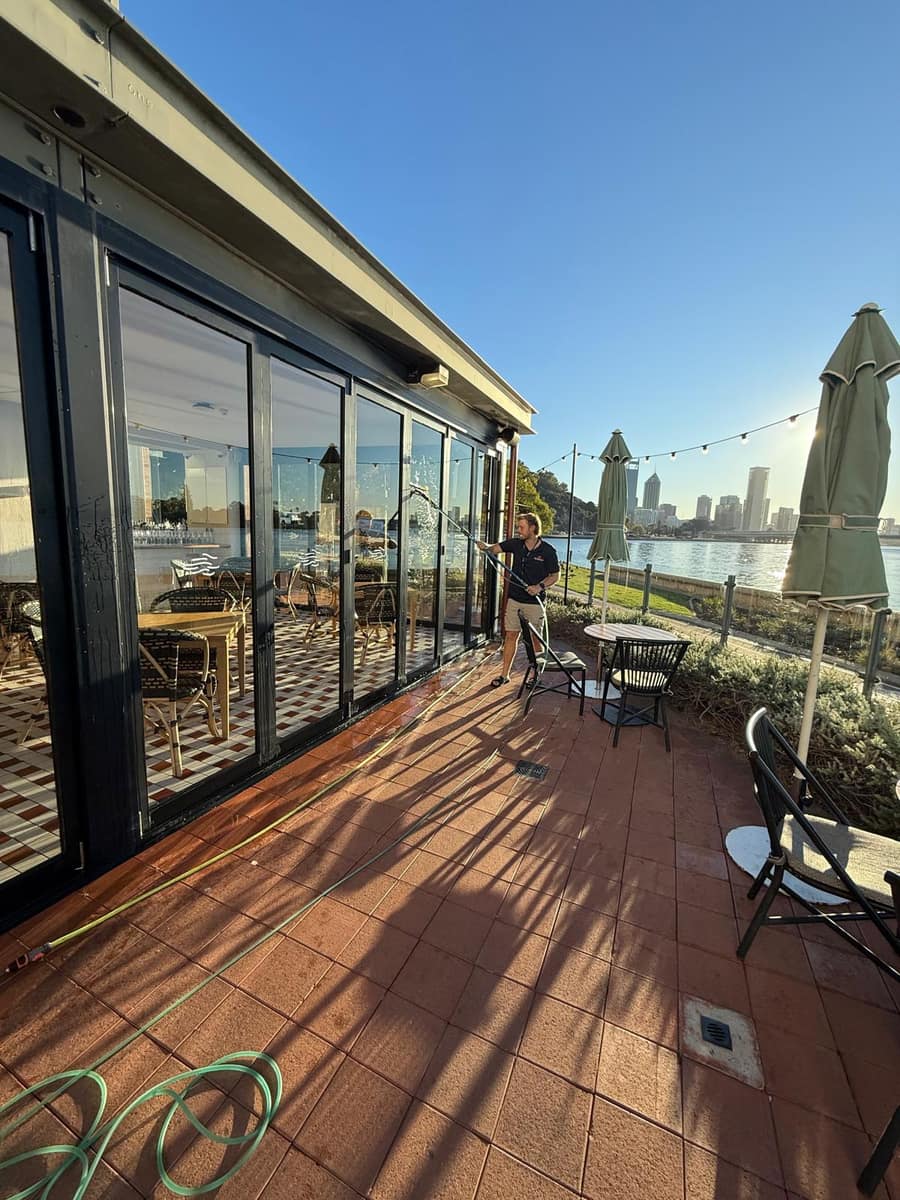 Worker cleaning glass bifold doors at a riverside venue, Perth skyline and Swan River in the background
